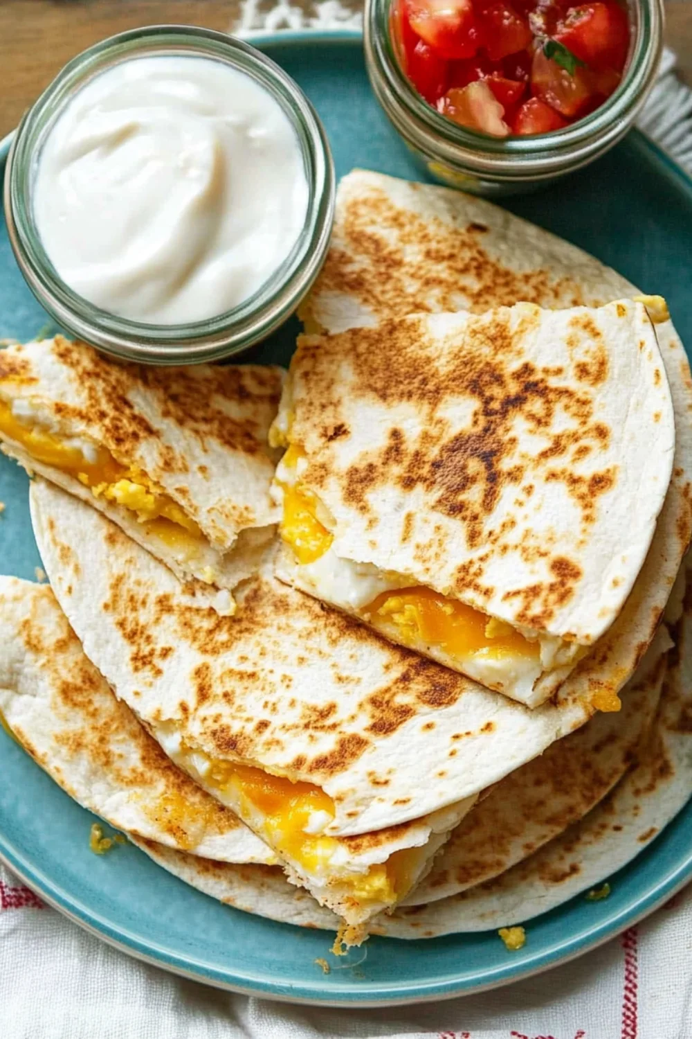 Golden brown egg quesadilla slices served on a teal plate with sides of sour cream and salsa.