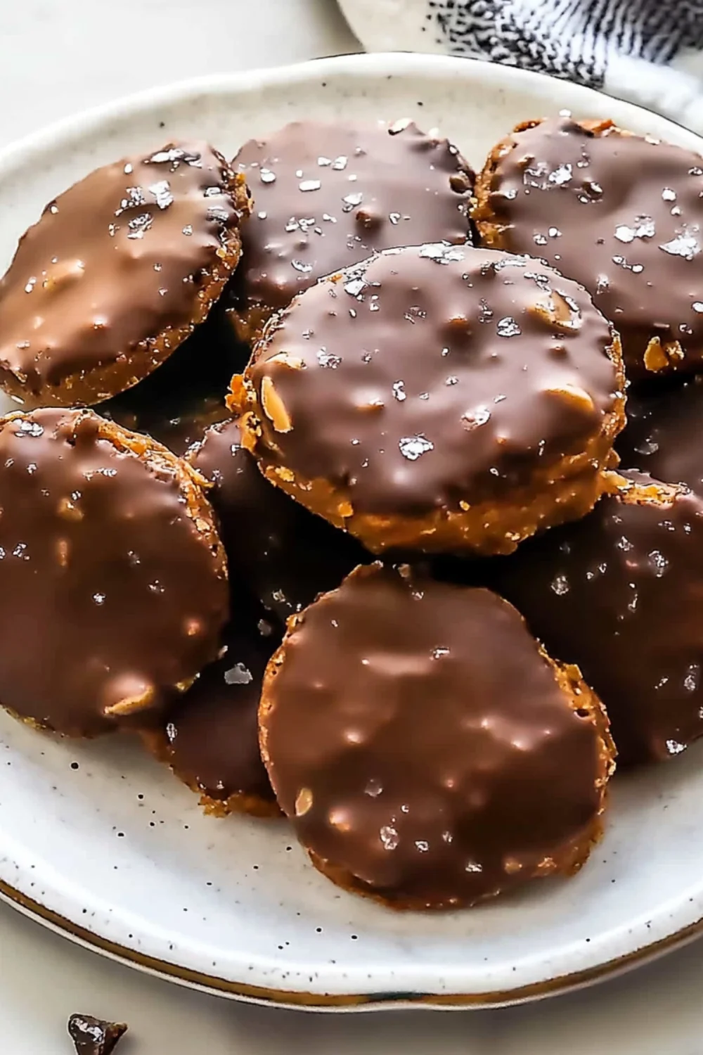 A close-up view of homemade date cookies coated in rich dark chocolate and sprinkled with sea salt, served on a ceramic plate.