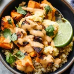 A close-up image of a bowl filled with roasted sweet potatoes, cauliflower, and quinoa, drizzled with a creamy sauce, garnished with cilantro leaves and a lime wedge.